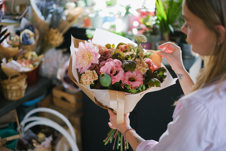 A woman with long blonde hair and a white shirt holds a vibrant bouquet of fresh flowers in a florist shop, with a variety of floral arrangements and supplies visible in the background. The bouquet fe