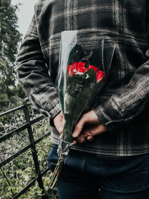 A person wearing a dark plaid jacket is holding a bouquet of fresh red and white roses wrapped in clear plastic with a black floral logo, positioned behind their back. The roses have vibrant petals an