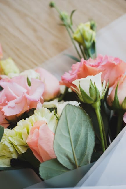 A close-up of a floral bouquet featuring soft pink lisianthus and pale yellow carnations, accented by green buds and large, velvety sage-green leaves. The flowers are arranged in a natural, slightly c