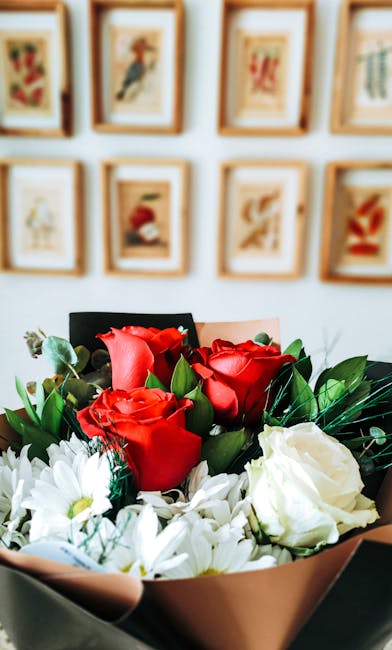 A close-up of a floral arrangement featuring vibrant red roses and white daisies, wrapped in light pink paper with green foliage accents. The bouquet is held in a person's hand, positioned in front of