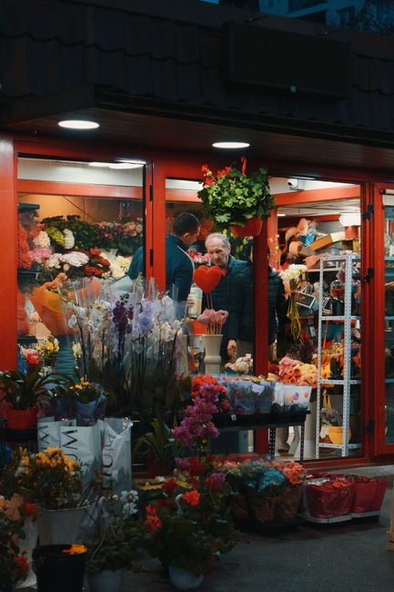 The image shows the exterior of a flower shop with a large glass window displaying an array of vibrant floral arrangements inside. Outside, a variety of potted plants and bouquets in different colours