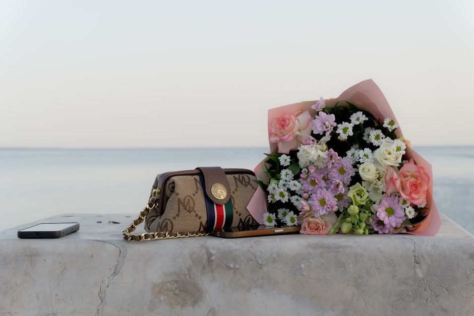 A bouquet of fresh flowers featuring pink roses, white daisies, and light purple chrysanthemums wrapped in soft pink paper is resting on a light-colored stone surface. Next to it, a designer handbag w
