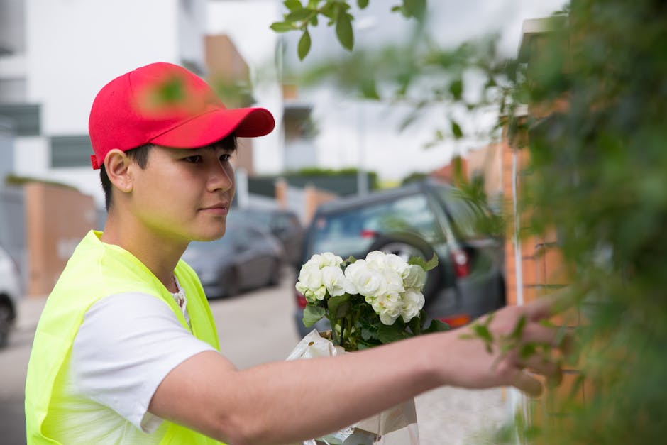 A young male florist wearing a red cap and a neon yellow vest is arranging a bouquet of white hydrangeas and roses on a porch or outdoor space. The arrangement is wrapped in white paper with a ribbon.